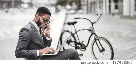 An executive black man is working on his laptop and speaking on his phone outdoors, with his bike nearby. The atmosphere is efficient yet relaxed, highlighting a balanced work-life approach. 117498805