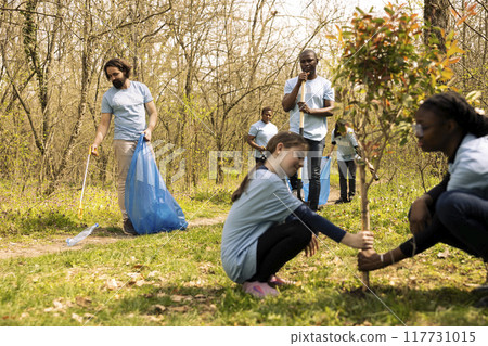 Diverse team of climate activists picking up garbage from the ground, collecting rubbish and recycling plastic in bags. People tidying the forest environment from trash, illegal dumping. 117731015