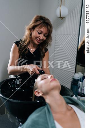 Vertical shot of pretty woman with blonde hair leaning on sink while smiling hairdresser washing hair in beauty salon. Beautiful female client feeling relax and happy while hairdresser wetting. 117755482