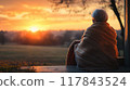 Elderly person enjoying a peaceful sunset wrapped in a blanket while sitting on a porch overlooking a serene landscape. Generative AI 117843524