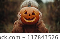 Young child happily holding a carved pumpkin mask outdoors during autumn near a rustic area in the late afternoon sunlight. Generative AI 117843526