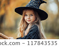 A young girl wearing a witch hat smiles while holding a broomstick in a forest during autumn. Generative AI 117843535