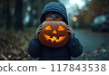 A child wearing a dark jacket holds a carved pumpkin featuring a smiling face on a quiet autumn evening. Generative AI 117843538