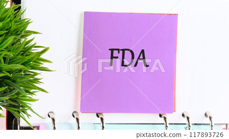 Food and Drug Administration (FDA) is a federal agency of the Department of Health and Human Services. FDA on a purple sticker on the background of a white leaf and a green plant in the foreground 117893726