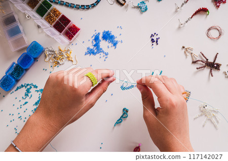 A girl's hands weave a beaded ring on a wire above a work table with finished jewelry 117142027