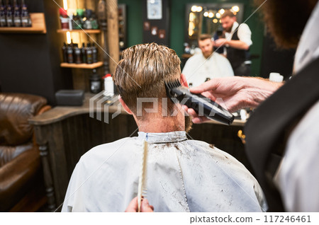 Professional barber using comb and electric shaver to cut side hair. Close-up view of male customer getting stylish haircut. 117246461