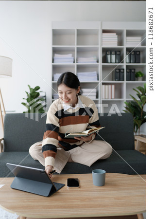 A woman sits on a sofa, using her notebook and tablet computer simultaneously to study online, efficiently managing her learning through digital platforms from the comfort of her home 118682671