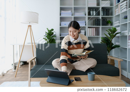 A woman sits on a sofa, using her notebook and tablet computer simultaneously to study online, efficiently managing her learning through digital platforms from the comfort of her home 118682672