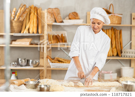 Woman works in bakery as baker, cuts dough into portions, forms pieces of dough to create buns. 118717821