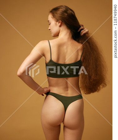 Rear view. Young woman with slim, fit body posing in underwear, showing her relieve back, smooth buttocks against sandy color studio background 118749940