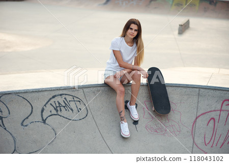 Young attractive girl in a white t-shirt and sneakers in modern skatepark with skateboard. 118043102