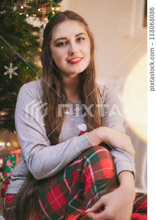 Vertical Portrait of young caucasian woman in festive pajama sits near Christmas tree in living room decorated for celebration of Christmas and New Year with garland lights bokeh. Cosy winter holiday. 118068086