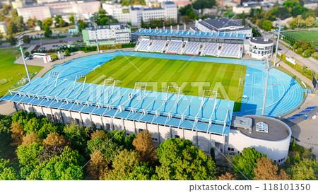 A high-quality aerial view of a modern sports stadium, featuring a football field and running track, surrounded by lush greenery and urban landscape. 118101350