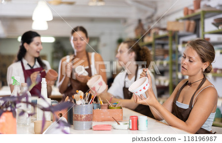 Young woman sits near table and decorates clay ceramic craft vase, plate 118196987