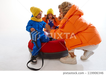 Mother dresser in bright orange winter jacket, preparing kids for snow tubing. Kids dressed in warm clothes and knitted hat, scarf and gloves. 119906346