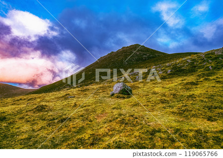 A mountain range with multiple peaks in the distance is partially obscured by clouds. The foreground features a grassy field with scattered rocks. 119065766