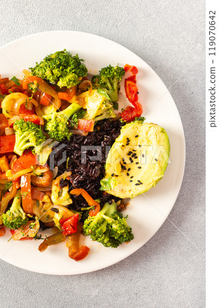 A hearty and nutritious breakfast of black rice with stewed vegetables like tomatoes, zucchini, peppers, and onions, fresh salad with tomatoes, broccoli, and radish, avocado 119070642