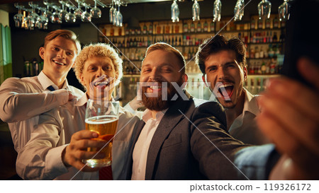 Business team gathers for selfie with smiles and beer glasses, capturing essence of fun, casual team outing in bar setting. 119326172