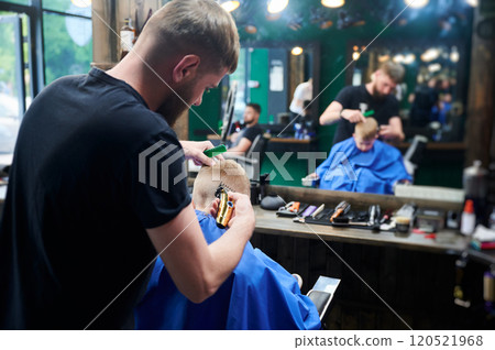 Hairdresser shaving boy's head. Competent barber using electric shaver at barbershop. Little client getting stylish men's haircut. 120521968