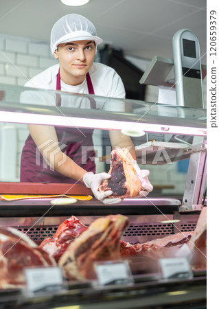 Positive young salesman demonstrating piece of meat in butcher shop 120659379