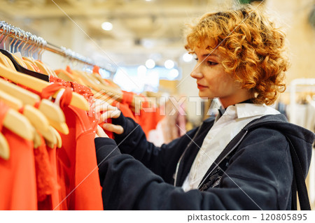 young attractive red-haired woman choosing red clothes in boutique 120805895