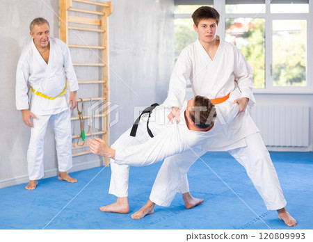 Two men practice grabbing and throwing sports mats during judo training under the guidance of an experienced mature coach 120809993