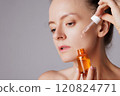 Happy smiling woman applying serum on her face, close eyes with pleasure. Redhead girl holding dropper with skin care product for healthy and glowing skin tone 120824771