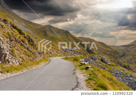 Winding mountain road through green slopes under a dramatic cloudy sky. 120992320