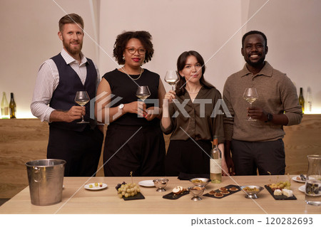 Croup portrait of multiracial professional sommeliers holding glass with white wine and smiling, wooden table with snack for wine in front of them 120282693