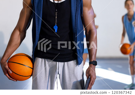 Holding basketball, coach with whistle standing in gym with players in background 120367342