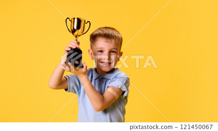 Happy boy raising gold trophy high, with medicine patch on arm, showing confidence and excitement against yellow studio background. 121450067