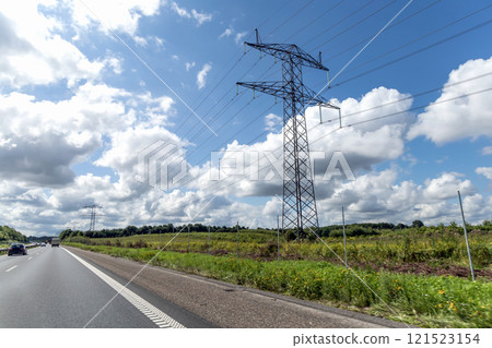 High voltage power line pylons, electrical tower on a green field with blue sky. Highway with cars on a cloudy summer day. 121523154