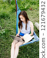 young woman with long hair and shorts sits in a hammock in the summer and works online on a laptop on the street. 121679432