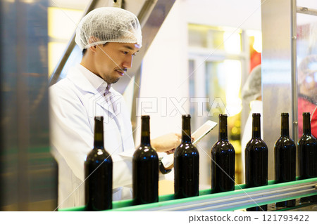 An empty glass bottles are rolling on the alcohol bottling belt machine in the manufacturing facility, meanwhile, male quality control supervisor is examining a bottles on the line. 121793422