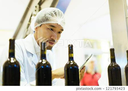 An empty glass bottles are rolling on the alcohol bottling belt machine in the manufacturing facility, meanwhile, male quality control supervisor is examining a bottles on the line. 121793423