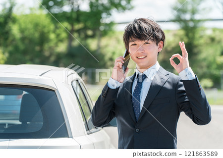 A young Asian businessman in a suit talking on a smartphone in front of a car 121863589