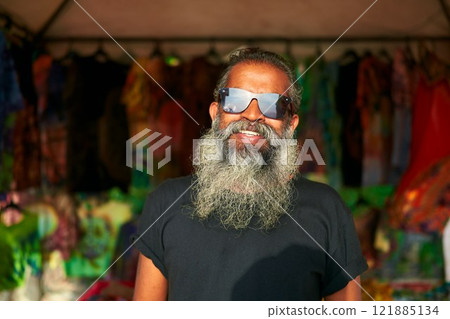 Senior artist with grey beard smiles at craft fair. Wears tie-dye t-shirt. Sunglasses reflect vibrant booths. Creative stall display. Daytime outdoor event. 121885134