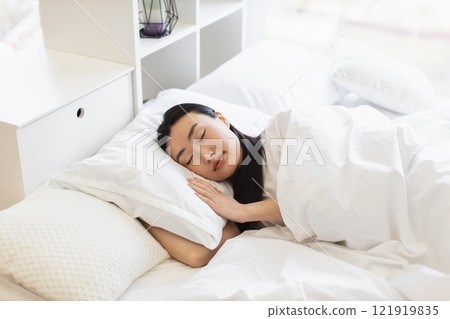 Asian woman enjoying peaceful sleep in white bed, showcasing relaxation and comfort. Serene morning atmosphere conveys sense of tranquility and rest. Ideal concept for sleep, rest, and wellness themes 121919835