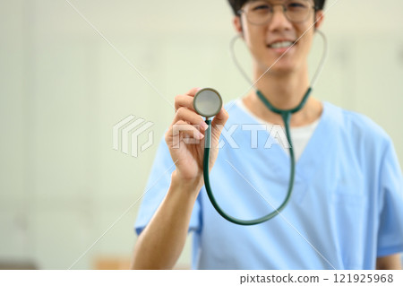 Smiling medical student in scrubs holding a stethoscope towards the camera 121925968