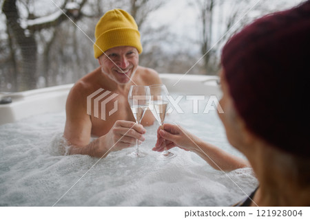 Senior couple bathing in hot tub during cold winter day, toasting with champagne. Romantic wellness getaway with spa during Valentine's weekend. 121928004