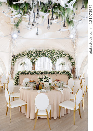 Elegant wedding reception hall featuring round tables, white and gold chairs, and floral arrangements 121973975