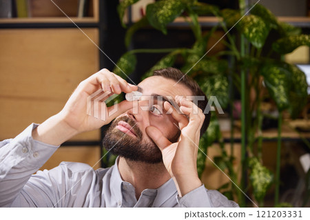 Man pulls down his eyelid and puts drops in his eyes to relieve dryness and irritation while sitting at desk at work. Disease and treatment. 121203331