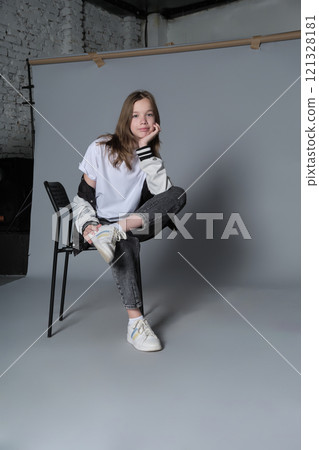 A girl sits with one arm resting on a chair, looking straight ahead. This portrait captures the spirit of modern youth, blending calm self-assurance with effortless style. 121328181