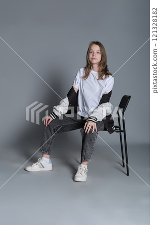 A teenage girl sits casually in a chair, her gaze steady and assured. Her posture reflects the self-confidence and ease of todays youth, embracing a minimalist style. 121328182