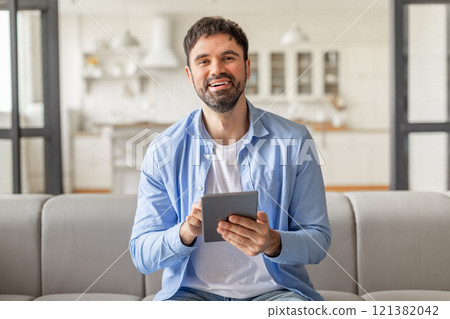 A man sits on a gray couch in his home and smiles while holding a tablet computer in his hands, websurfing or scrolling 121382042