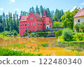 Cervena Lhota Castle rises majestically from a rocky island, surrounded by a serene lake and lush greenery. The vibrant red structure contrasts beautifully with the natural landscape. 122480340