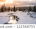 Snow blankets the landscape around Jizerka Stream in the Jizera Mountains, creating a tranquil winter scene. Tall evergreens and a golden sunset enhance the serene atmosphere. 122568755