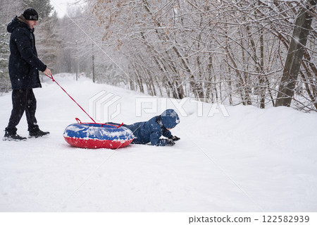 a child flips over from snow tubing while riding with his dad against the backdrop of a winter forest. Dangerous skiing in winter. Children's injuries during winter sports 122582939