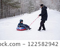 dad in winter jacket rides child on snow tubing around himself, against the background of snowfall and forest in winter. Dangerous and fun games of dad and son in winter 122582942