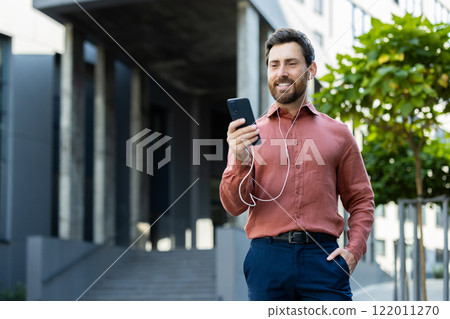 Businessman using wired headphones talking to colleagues and partners while walking around the city. Man smiling contentedly outside office building. 122011270